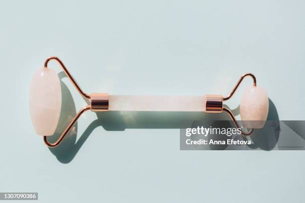 rose quartz roller for facial massage on pastel light blue teal background. sunlight makes shadows and illuminating reflections from tool's metallic part. massagers for anti aging procedures with an oil or moisturizer. flat lay style with copy space - anti mask fotografías e imágenes de stock