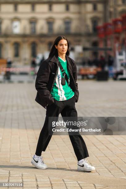 Model wears a a black jacket, a green and white stripe v-neck soccer t-shirt, a mini fanny pack bag crossbody, black pants, white socks, white Nike...