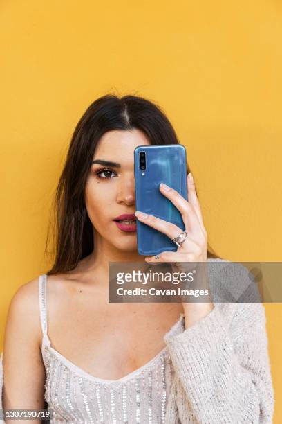 portrait of spanish brunette girl covering one eye with her mobile phone on yellow background wall. - coprire foto e immagini stock