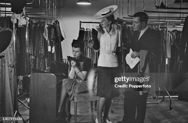 English fashion designer Norman Hartnell works with two female fashion models on final adjustments to a Utility clothing fashion range at his salon...