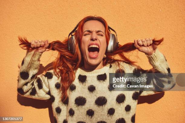 white caucasian woman with red hair wearing helmets to protect herself from noise pulls her pigtails while screaming. noise concept - reduce stock pictures, royalty-free photos & images