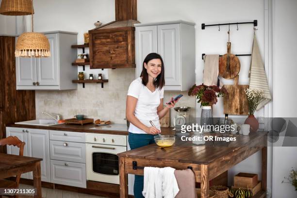 happy woman with smartphone preparing cake in kitchen at home - cake bowl stock pictures, royalty-free photos & images