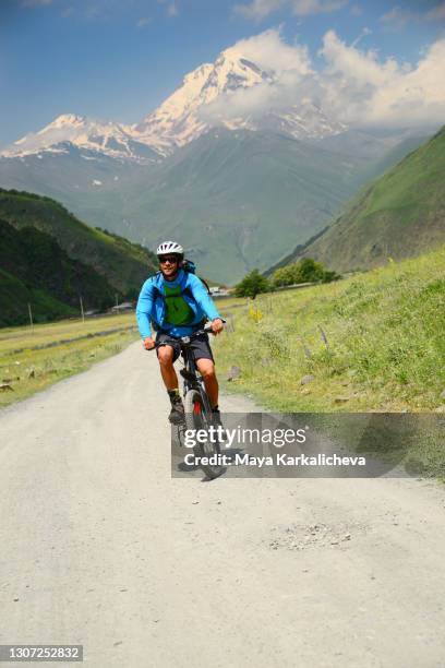 young man riding a bicycle on a scenic mountain road in a sunny summer day.snowcapped mount kazbek in the background ( 5054m altitude ) situated in caucasus mountain - mountain biker stock-fotos und bilder