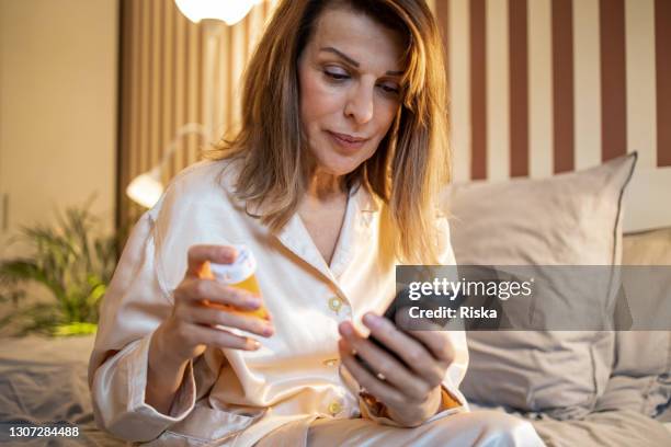 rijpe vrouw in de slaapkamer, die de telefoon bekijkt en een pilfles houdt - slaappil stockfoto's en -beelden