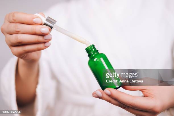 woman in white bathrobe with nude pink manicure holds pipette and bright green glass bottle with natural moisturizer or organic serum. she ready to start skin care procedures. concept of home body care and healthy lifestyle. close-up front view - ホホバ ストックフォトと画像