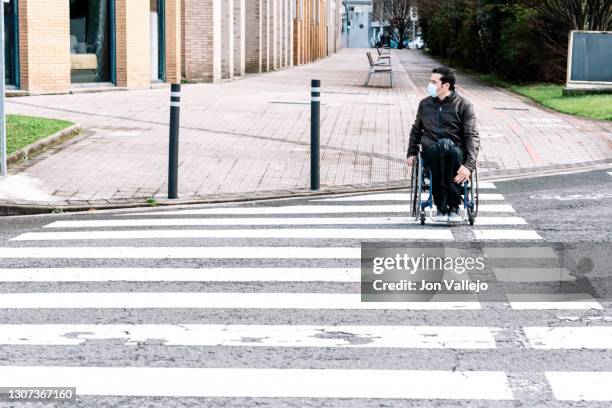 handicapped man with mask facing right as he crosses a crosswalk in his wheelchair. - zebra crossing stock-fotos und bilder