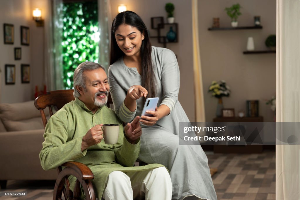 Portrait of a young woman and her mature father sitting on rocking chair at home. she is showing something in mobile phone :- stock photo