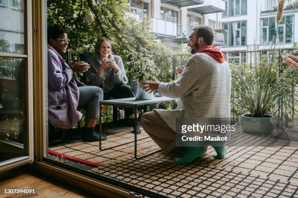 young man spraying plant while looking at female friends in patio - cultural diversity stock-fotos und bilder