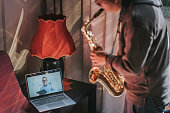 Lifehack asian chinese mid adult man practicing saxophone at living room online virtual class with his tutor using laptop
