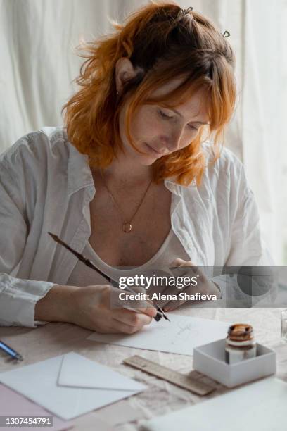 a calm red-haired woman sits at a table with a pink linen tablecloth and is engaged in calligraphy, writing cards with pen and ink. hobbies, meditation, mindfulness, homework. - caligrafia imagens e fotografias de stock