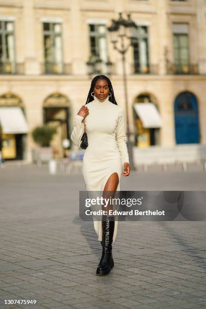 Magalie Kab wears earrings, a white wool turtleneck long slit dress, a black leather bag with pearls, black leather knee-high combat boots, on March...