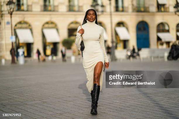 Magalie Kab wears earrings, a white wool turtleneck long slit dress, a black leather bag with pearls, black leather knee-high combat boots, on March...