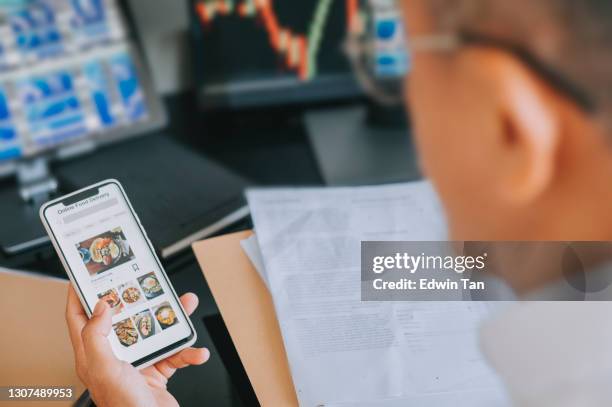 online food delivery mobile app shown on smart phone screen hold by asian man hands in front of desktop pc - dinner program stock pictures, royalty-free photos & images