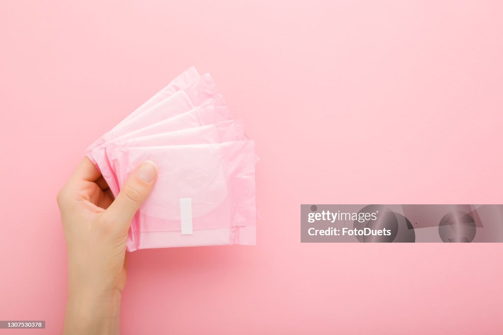 Young adult woman hand holding packs of sanitary towel on light pink table background. Pastel color. Closeup. Empty place for text. Top down view.