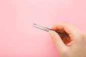 Young adult woman fingers holding tweezers on light pink background. Pastel color. Closeup. Top down view.