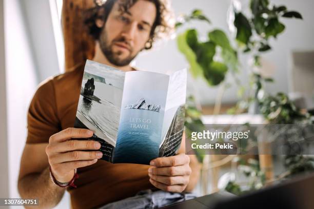 young casually clothed man looking at travel flyer in his apartment - flyer leaflet stock pictures, royalty-free photos & images