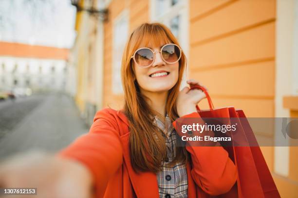 self portrait of redhead girl in modern coat with red shopping bags in the city - red sunglasses stock pictures, royalty-free photos & images