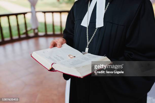 religious ceremony of marriage, a catholic priest in black clothes holding a bible in red covers, two gold rings between the pages, outdoors, summer, green grass, close up - priest stockfoto's en -beelden