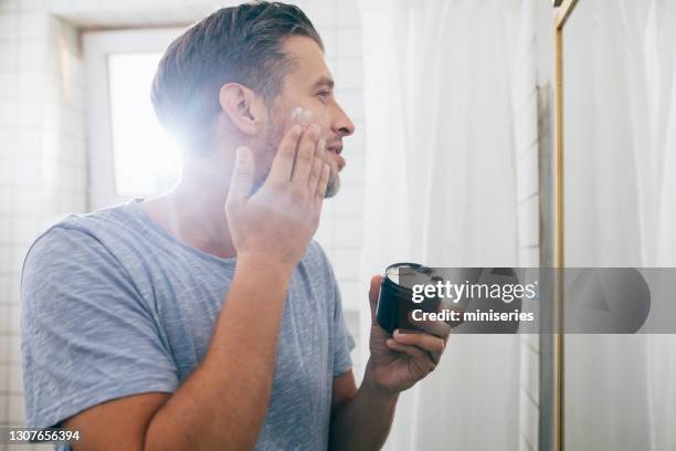 handsome young man applying face cream after a morning shave - mens cologne stock pictures, royalty-free photos & images