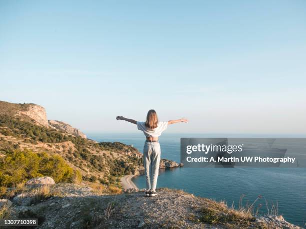 young woman with arms wide open standing on rock and looks to ocean - girl hair stock pictures, royalty-free photos & images