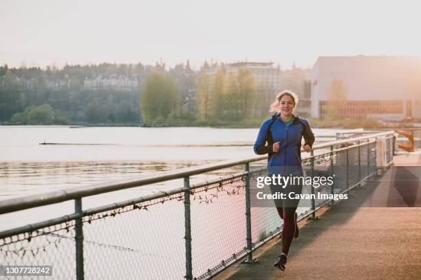 woman run next to lake view with warm sunshine glowing behind her - carrera de carretera fotografías e imágenes de stock