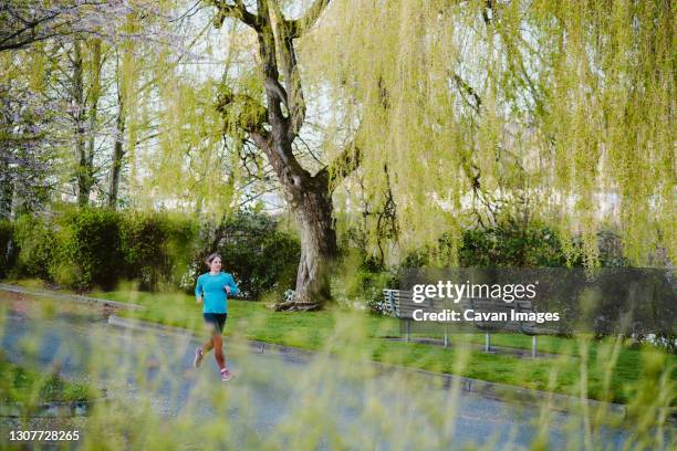 an athletic woman runs in city park surrounded by wispy green trees - maillot de sport stockfoto's en -beelden