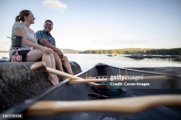 african american man and white woman sit on rock on lake with canoe - two people canoeing on a lake stock pictures, royalty-free photos & images