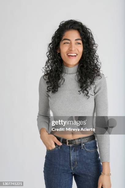 portrait of a latinx millennial woman with curly hair, stands in front of a white backdrop in a studio setting, while wearing a grey cropped top and blue jeans. - bauchfreies oberteil stock-fotos und bilder