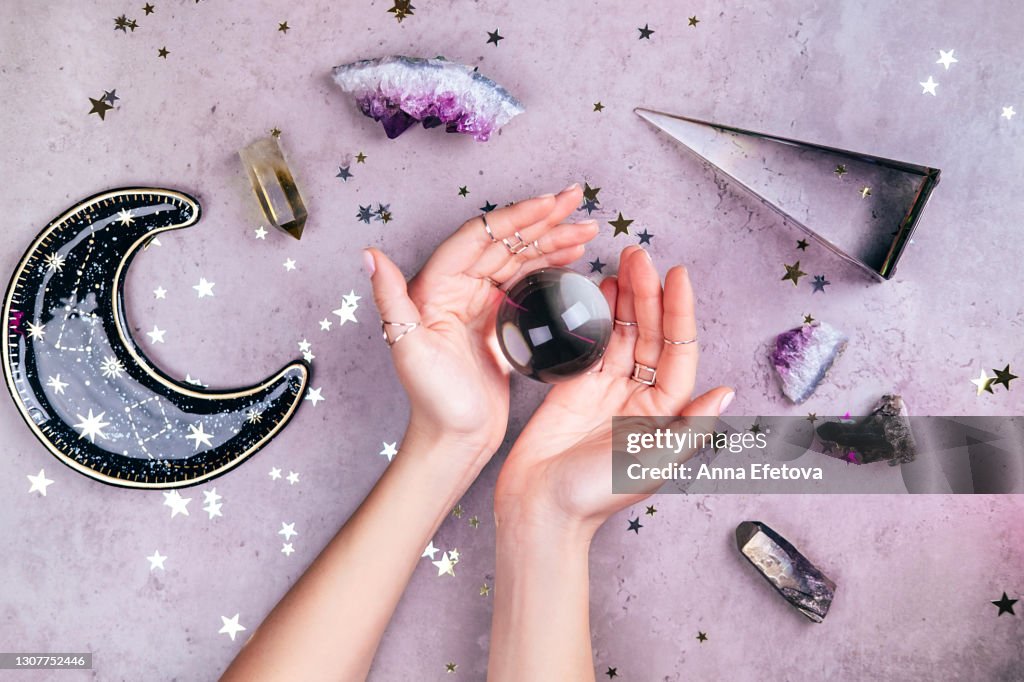 Magic crystals for rituals. Hands with rings on fingers are holding crystal ball near esoteric set on concrete gray background with many stars sequins. Flat lay style