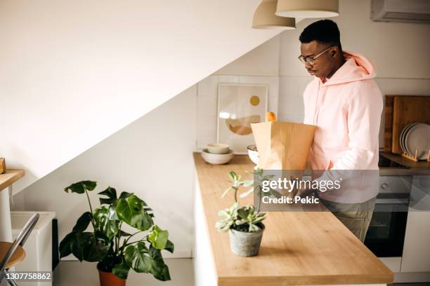 young man unpacking his grocery paper bag at home - young man groceries kitchen stock pictures, royalty-free photos & images