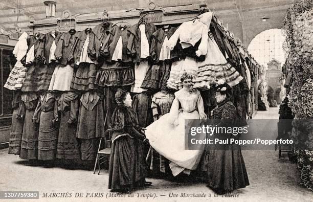 Postcard showing two women inspecting a mannequin at a dress stall in the Marches De Paris, the women wear dark bustle skirts and velvet bolero...