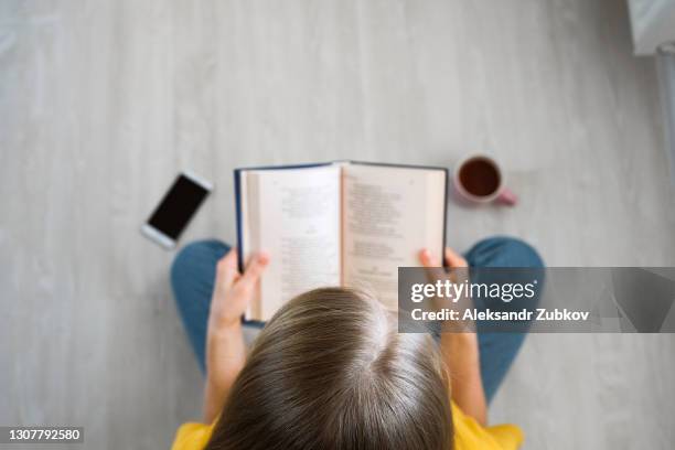 the girl is sitting on the floor with her legs crossed in the lotus position and reading a book. a woman holds a textbook in her hands. next to it is a mug of tea or coffee and a mobile phone. the concept of education, development and self-improvement. - poetry literature stock pictures, royalty-free photos & images