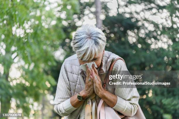 de hogere vrouw met griep loopt in openbaar park en niezend in document weefsels. - hooikoorts stockfoto's en -beelden