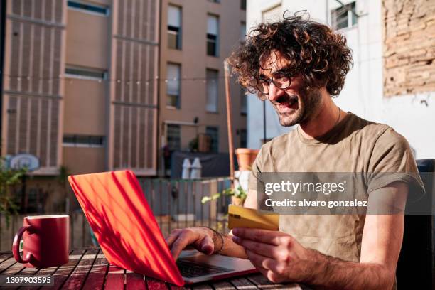 a young man buying online with his credit card and laptop at his home in a sunny day - kundenzufriedenheit stock-fotos und bilder