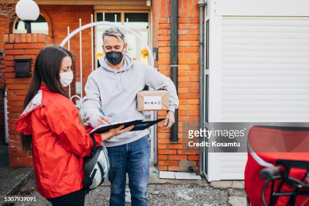 mannelijke klant, die het document over het ontvangen van leveringspakket ondertekent, terwijl allebei beschermend gezichtsmasker dragen tijdens covid-19 pandemieuitbraak als nieuw normaal - postbode stockfoto's en -beelden