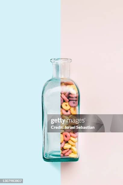 close up of a composition of a empty and full glass bottle with coloured ring shaped cereals inside on a light pink and blue background. concept of minimalism - moitié photos et images de collection