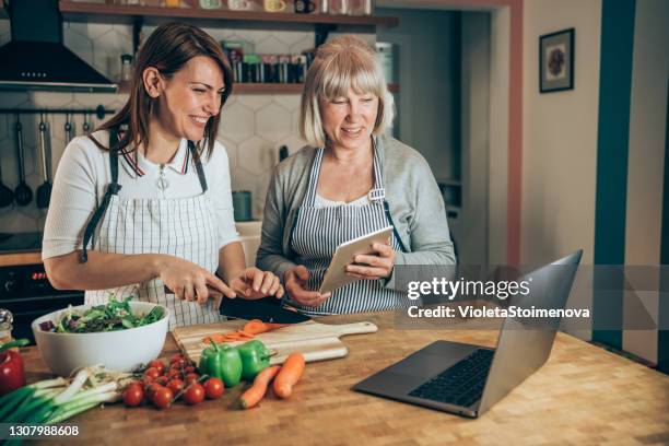 mère et descendant utilisant l’ordinateur portatif tout en cuisinant le repas sain dans la cuisine. - cours de cuisine photos et images de collection