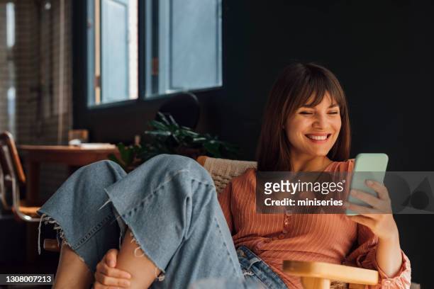 frauen in der wirtschaft: schöne lächelnde junge geschäftsfrau mit einem handy, während sie lässig in einem stuhl sitzt - smartphone zuhause stock-fotos und bilder