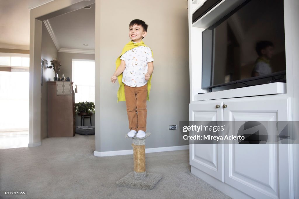 A young mixed race toddler with short hair, excitedly stands on a cat scratching post wearing a green towel around his neck as a cape and tan colored pants with a white patterned shirt.