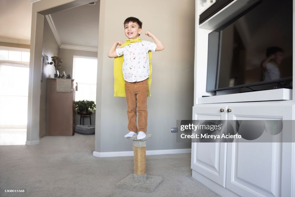 A young mixed race toddler with short hair, excitedly stands on a cat scratching post wearing a green towel around his neck as a cape and tan colored pants with a white patterned shirt.