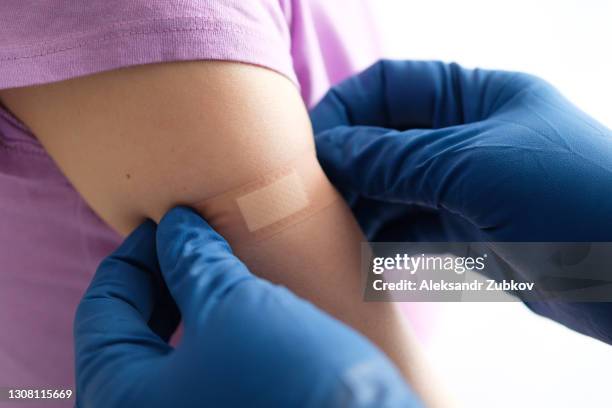 a gloved doctor or health care professional applies a patch or adhesive bandage to a girl or young woman after vaccination or drug injection. the concept of medicine and health care, vaccination and treatment of diseases. first aid services. - vaccination foto e immagini stock