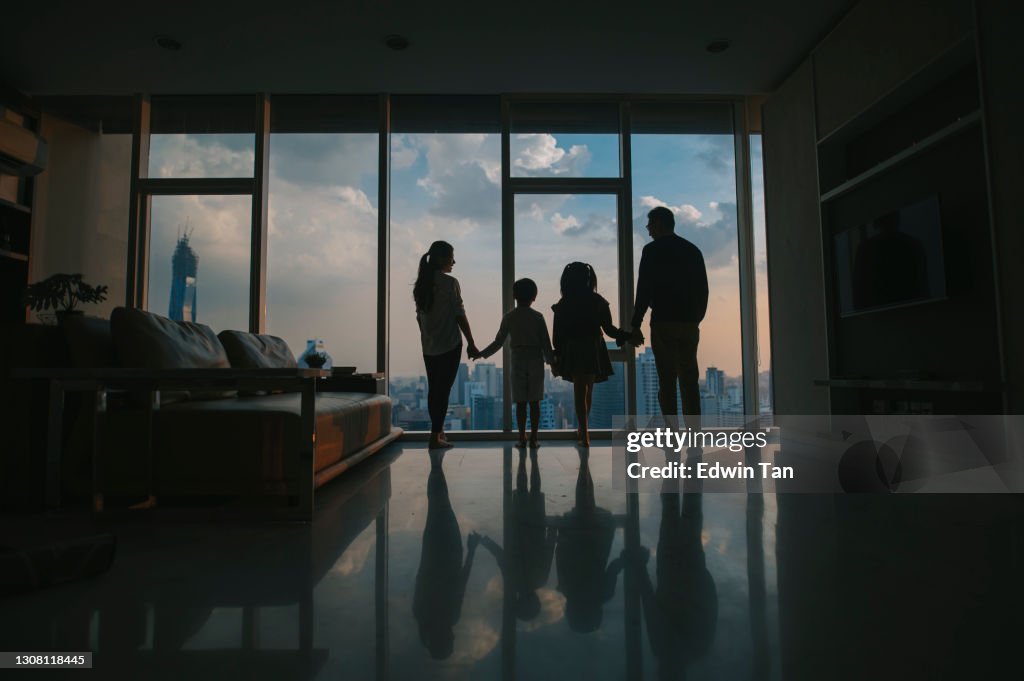 Silhouette rear view  asian chinese family with 2 children looking through window enjoying beautiful sunset at living room in kuala lumpur city