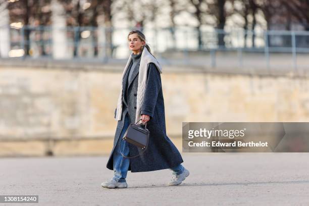 Xenia Adonts wears golden earrings, a gray wool turtleneck pullover, a white knit wool oversized pullover, a gray long oversized wool coat, blue...