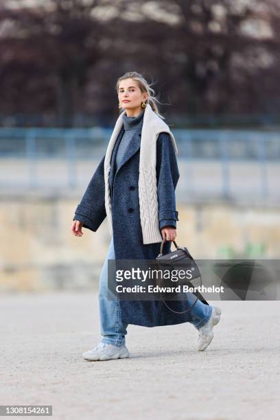 Xenia Adonts wears golden earrings, a gray wool turtleneck pullover, a white knit wool oversized pullover, a gray long oversized wool coat, blue...