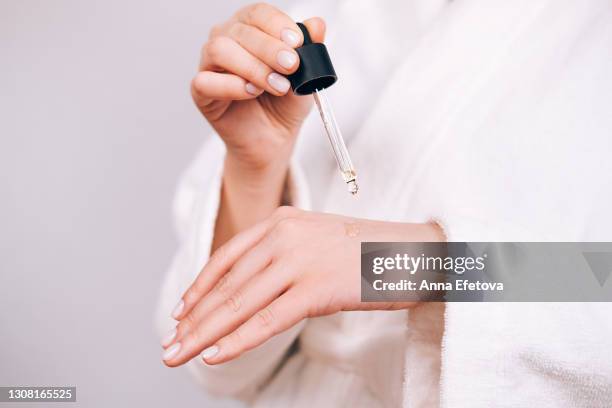 woman in white bathrobe with nude pink manicure holds glass pipette with natural essential oil or organic serum. moisturizing oil is dropping on hand's skin from pipette. concept of home body care and healthy lifestyle. close-up front view - ホホバ ストックフォトと画像