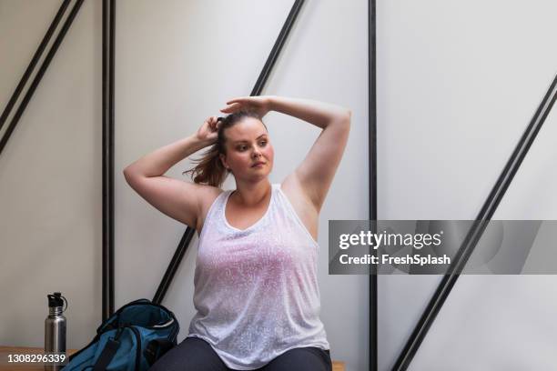 mooie plus size vrouw in sportkleding zitten in de kleedkamer en binden haar haar in een paardenstaart - maillot de sport stockfoto's en -beelden