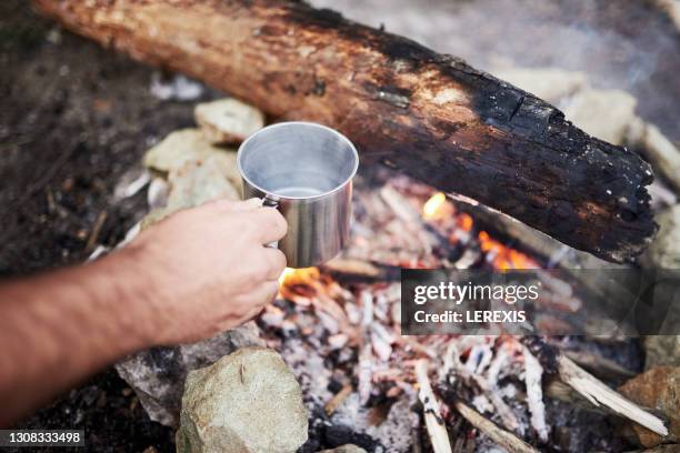 hand holding a metal cup over a fire lit in a camping in nature - emaille stock-fotos und bilder