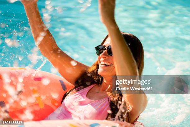 cheerful woman with inflatable ring splashing water while enjoying in swimming pool - water glasses ストックフォトと画像