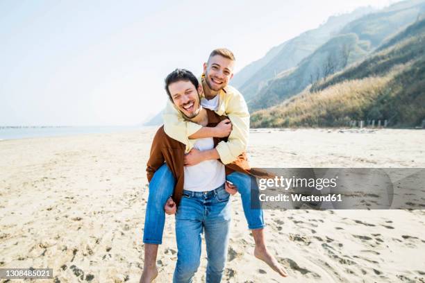 cheerful gay man giving piggyback to friend on beach during vacations - piggyback stock pictures, royalty-free photos & images