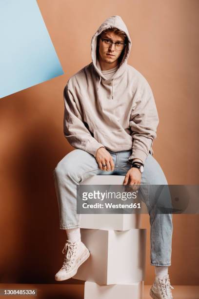 serious man in casual clothing sitting on box container against brown background - kapuzenoberteil stock-fotos und bilder